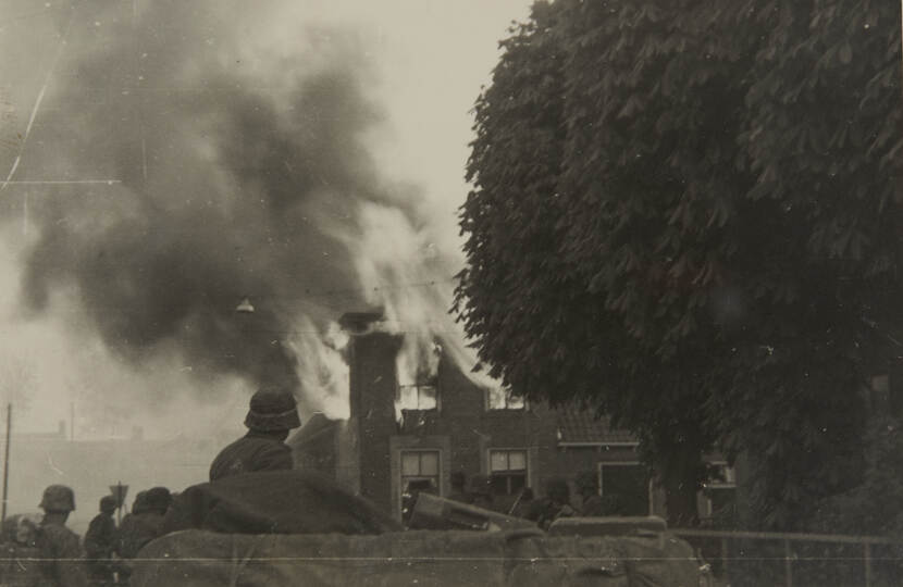 Zwart-witfoto. Enkele militairen staan naar een huis dat in brand staat te kijken, hoge vlammen schieten door het dak en ramen naar buiten, dikke rook hangt boven straat. Rechts een grote boom.