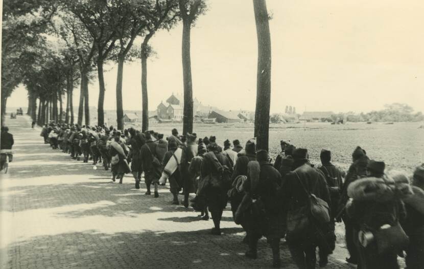 Zwart-witfoto. Verharde weg met bomen aan weerszijden, aan de rechtskant lopen tientallen militairen in een lange rij met drie of vier naast elkaar. De militairen dragen lange jassen en mutsen, velen dragen rugzakken, tassen en dekens op hun rug of over hun schouders. In de achtergrond bebouwing van een dorp en akkerland.