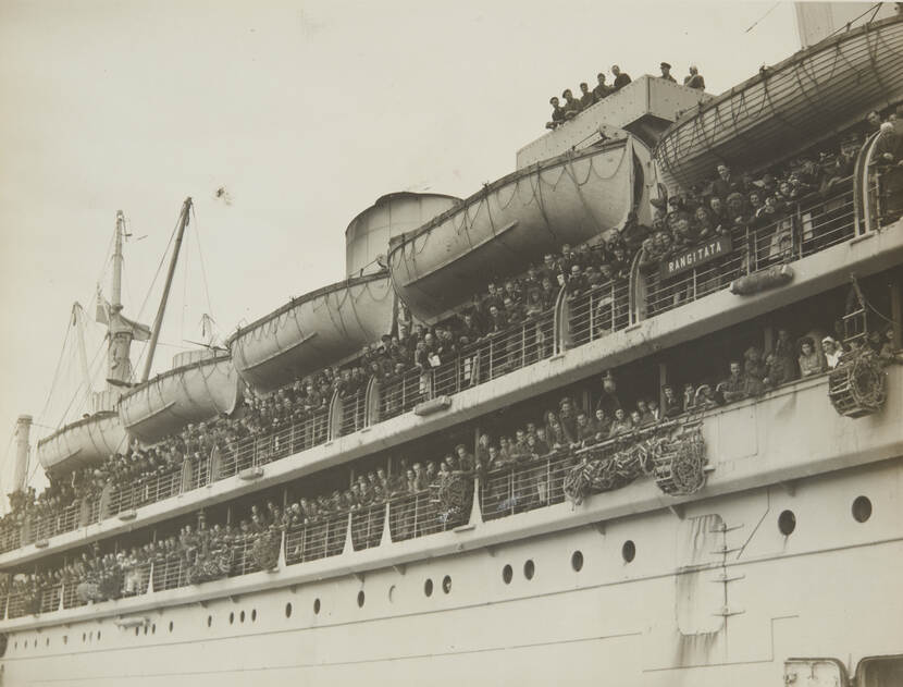 Zwart-witfoto van de zijkant van een transportschip, veel mensen hangen over reling te zwaaien.