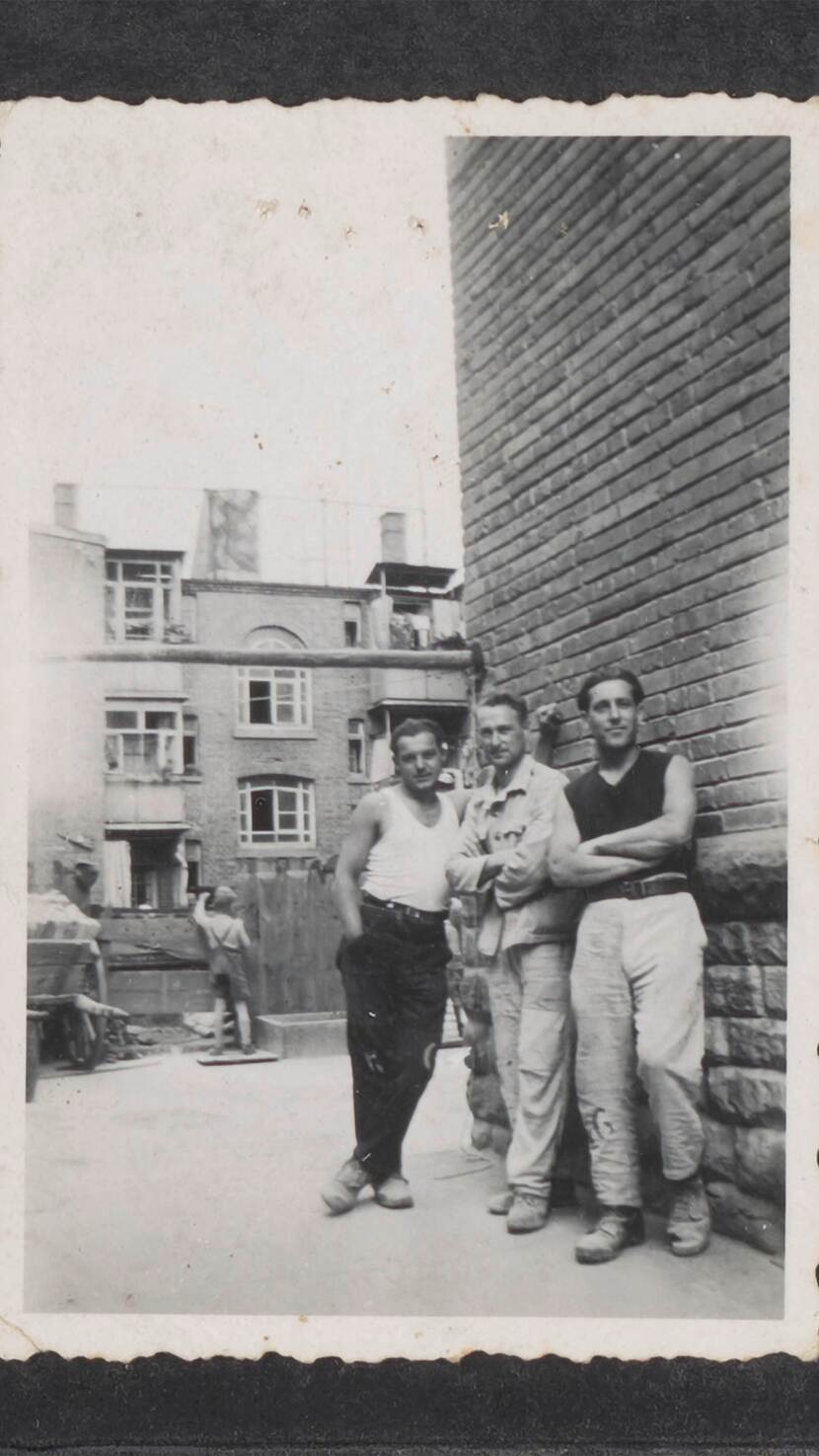 Drie mannen poseren voor de foto. Zij staan voor een muur en schuin op een rijtje. Op de achtergrond zijn wat huizen te zien.