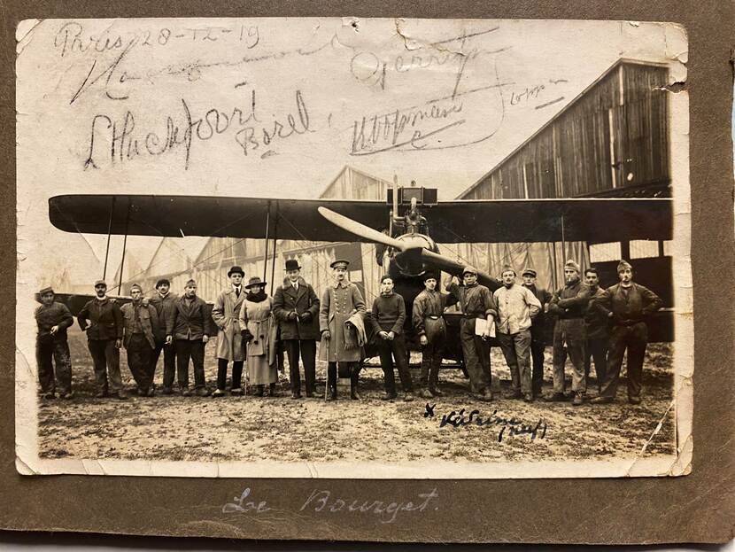 Zwart-witfoto geplakt in een fotoalbum, van een groep mensen voor een dubbeldeks propellervliegtuig.