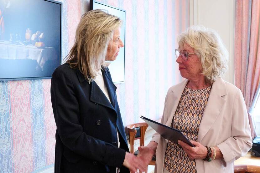 Kleurenfoto van Minister van Defensie Kajsa Ollongren (links) die de hand schud van een oudere vrouw, de dochter van marinier Joop de Hoog. De dochter heeft een map vast waar een brief in zit.