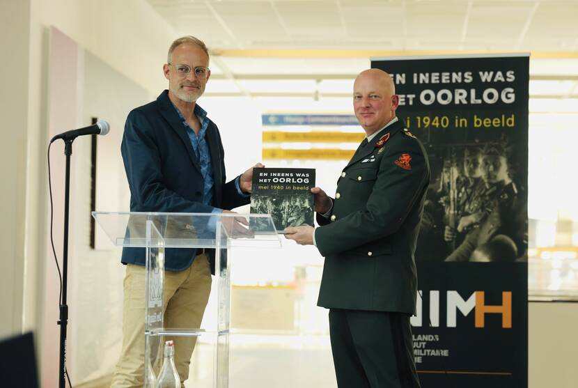 Op de foto wordt het boek door NIMH-medewerker overhandigd aan generaal-majoor Roy Sillen. Op de achtergrond is een NIMH-banner te zien.