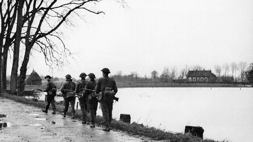 Militairen lopen links langs het water. Zij lopen achter elkaar.